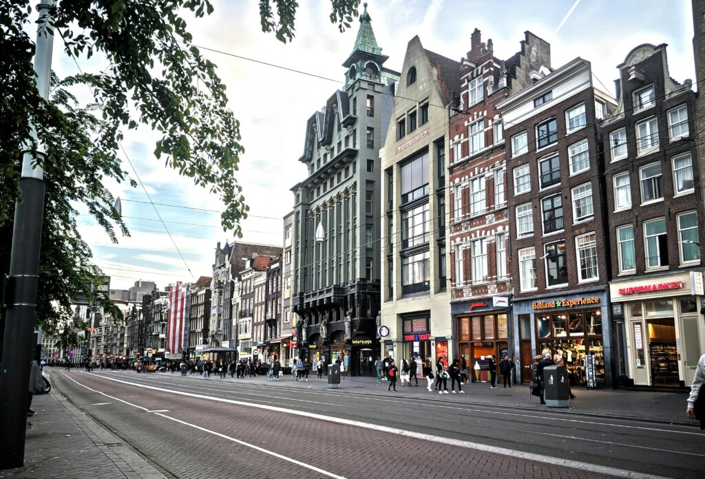 Busy Amsterdam city street with tall Dutch canal buildings, tram lines, and pedestrians — UK to Netherlands removals to Amsterdam by Trunk Logistics