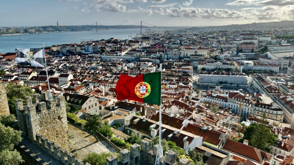 View across Lisbon from São Jorge Castle battlements with the Portuguese flag flying and the 25 de Abril Bridge over the Tagus in the distance — UK to Portugal removals to Lisbon by Trunk Logistics