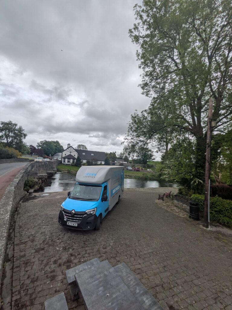 Trunk Logistics removal van beside an Irish river in County Tipperary with stone bridge and village visible — Ireland to UK removals from County Tipperary