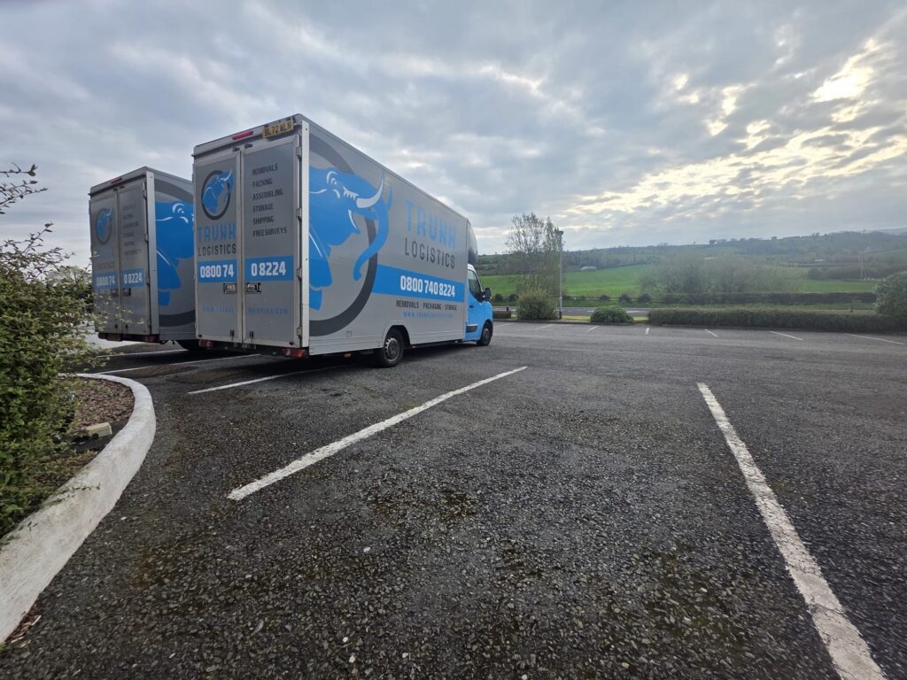 Two Trunk Logistics removal vans parked in Omagh, County Tyrone, Northern Ireland, with green countryside in the background
